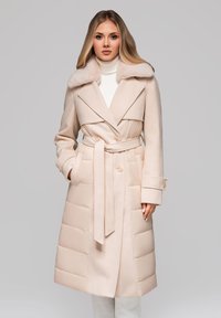 Žena v béžovém zimním kabátu s páskem, kožešinovým límcem a prošívanými panely, stojící na jednobarevném šedém pozadí.