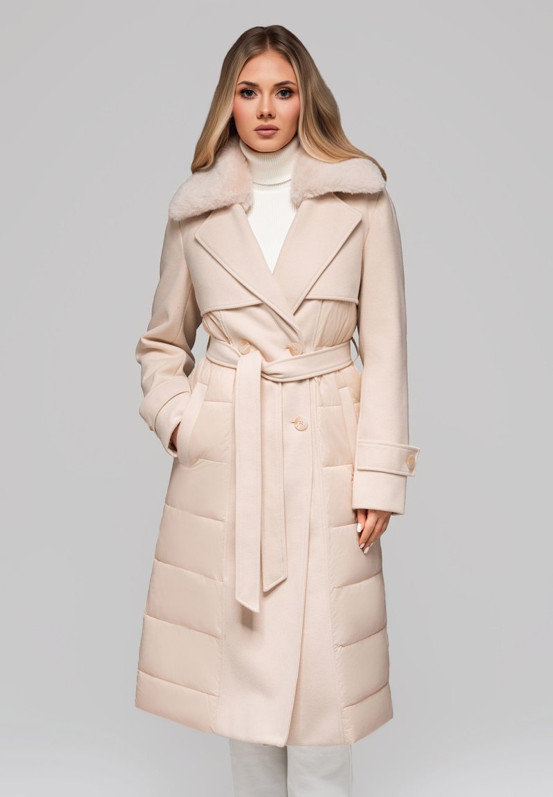 Žena v béžovém zimním kabátu s páskem, kožešinovým límcem a prošívanými panely, stojící na jednobarevném šedém pozadí.