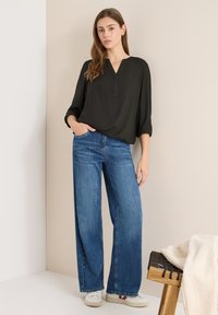 Schwarze, locker sitzende Bluse mit V-Ausschnitt und hochgekrempelten Ärmeln, kombiniert mit weit geschnittenen blauen Denim-Jeans und weißen Sneakern. Dezente Texturen.