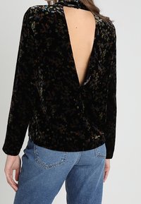 Sort velourbluse med blomstermønster, lange ærmer og dyb V-rygdesign. Båret med højtaljede blå denimjeans.