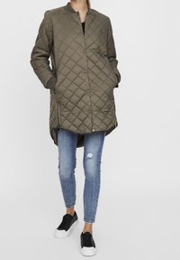 Oliivivihreä tikkikangas pitkä takki, jossa on napit, ribatut hihansuut ja sivutaskut, yhdistettynä kuluneisiin sinisiin farkkuihin ja mustiin lenkkareihin.