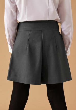 Bagudvendt udsigt af en person iført mørkegrå plisserede shorts over sorte strømpebukser og en hvid langærmet skjorte mod en beige baggrund.