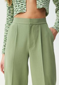 Pantalon vert plissé en tissu texturé, taille haute, avec poches latérales. Associé à un haut court en maille à motifs.
