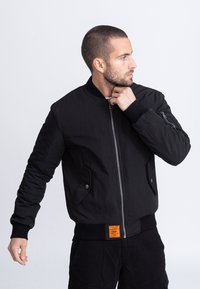 Giacca bomber nera con zip frontale, polsini a costine, due tasche frontali e un'etichetta arancione sull'orlo. Tessuto liscio e design aderente.