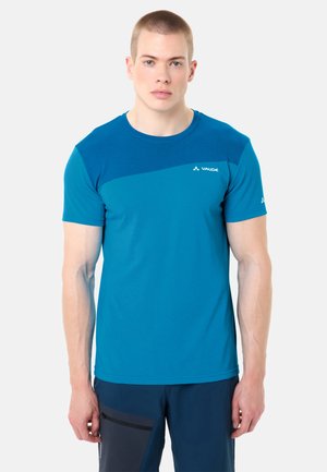 Junger Mann mit kurzen Haaren, der ein blaues Vaude-Sportshirt mit kurzen Ärmeln und dunkelblaue Hosen trägt, vor einem weißen Hintergrund stehend.