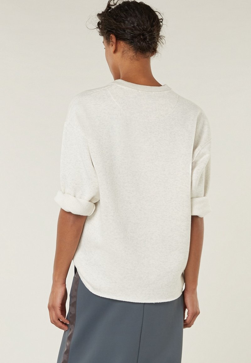 Personne aux cheveux courts et bouclés porte une chemise à manches longues gris clair avec les manches retroussées et une jupe sombre avec une bande latérale, vue de dos.