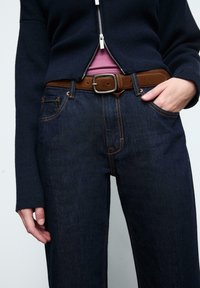 Tmavomodré denim džínsy s oranžovým šitím, v kombinácii s hnedým koženým opaskom a námorníckym pleteným topom, vytvárajúce ležérny, vrstvený vzhľad.