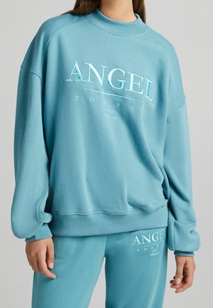 Osoba nosząca jasnoniebieską bluzę i pasujące spodnie z napisami "ANGEL ENERGY" i "JUST FEEL IT" na obu częściach.