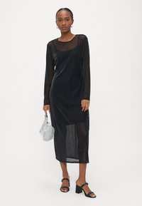 Μαύρο, διαφανές φόρεμα με μακριά μανίκια, στρογγυλή λαιμόκοψη και μήκος μέχρι τη μέση της γάμπας. Φορεμένο με σανδάλια με χοντρό τακούνι και ασημί μεταλλικό τσαντάκι χειρός.