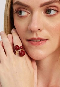 Zlato obarvana prstan s tremi rdečimi sijajnimi biseri različnih velikosti, nameščen na ozki obroček, nosi se na roki blizu obraza.