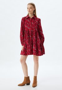 Rotes Paisleymuster-Kleid mit langen Ärmeln, Kragen, Knopfleiste und gestuftem Rock. Kombiniert mit braunen Wildleder-Stiefeletten.