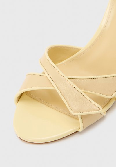 Μπεζ σανδάλι ανοιχτού δαχτύλου με δύο φαρδιές, λείες δερμάτινες λωρίδες που διασταυρώνονται εμπρός και σχέδιο προσαρμοσμένο σε τετράγωνο σχήμα στα δάχτυλα.