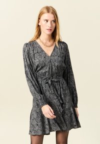 Schwarzes Kleid mit langen Puffärmeln, einem tiefen V-Ausschnitt und einem elastischen Taillenbund. Es verfügt über ein florales Muster und ein weiches, strukturiertes Material.