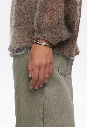 Pulsera trenzada de plata y oro en una mano, combinada con un suéter marrón suave y pantalones verdes. Textura suave y acabado metálico pulido.