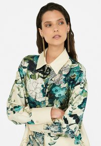 Bluse mit Blumenmuster, die einen cremefarbenen Grund mit grünen und blauen Mustern hat, einen spitzen Kragen und Knopfdetails an der Vorderseite und den Manschetten.