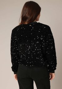 Bomber noir en sequins avec une finition texturée. Présente des poignets et un ourlet côtelés, mettant en valeur une surface brillante et réfléchissante.