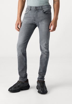 Mężczyzna ubrany w wyblakłe szare wąskie jeansy i czarne buty sportowe, stojący na tle jasnego, jednolitego tła.