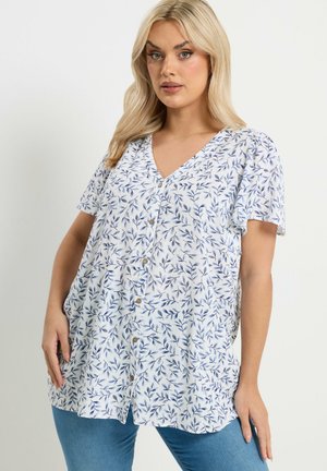 Blonde vrouw die een witte blouse met knopen draagt met een blauw bladpatroon en een blauwe spijkerbroek, staand met één hand in de broekzak.