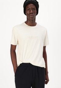 Kratke rokave, kremna majica s pisanjem "Hollister" iz vezenih svetlih niti. Uparjena z temno modrimi hlačami. Gladka tekstura in sproščen kroj.