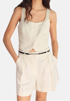 Kvinde iført ærmeløs beige cropped top med knapper foran og matchende høj talje shorts med sort bælte, hænderne i lommerne.