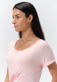 Camiseta de manga corta rosa claro con escote redondo y mangas enrolladas. Presenta un gráfico de hoja de palma en rosa más oscuro en la esquina inferior izquierda.