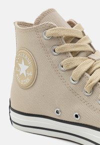 Bež Converse All Star visoke tenisice s teksturiranim platnenim materijalom, okruglim prstima, sistemom vezanja i bijelom gumenom petom s crnom prugom.