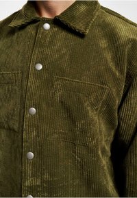 Olive green corduroy shirt jacket featuring vertical ribbing, silver snap buttons, and two chest pockets. Textured fabric gives a soft appearance.