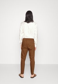 Bílá košile s dlouhým rukávem s tištěným vzorem, kombinovaná s hnědými zúženými kalhotami; outfitu dominuje casual obuv.