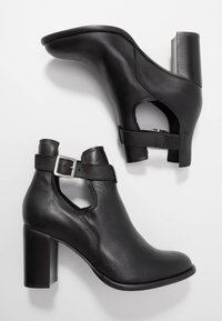 Bottines en cuir noir avec un bout arrondi, des côtés découpés et une bride à boucle. Les talons sont de forme carrée et mesurent environ 8 cm de hauteur.