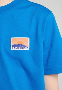 Sininen puuvillapaita, jossa on ommeltu etiketti, jossa on auringonlaskun kuvasto, siluettina vuoria ja sana "CALIFORNIA" sinisin ja oranssein värein.