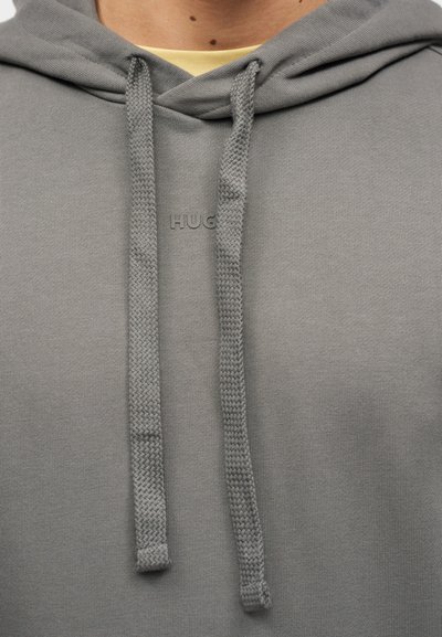 Gros plan sur une personne portant un sweat à capuche gris avec de gros cordons et le mot "HUGO" embossé sur la poitrine.