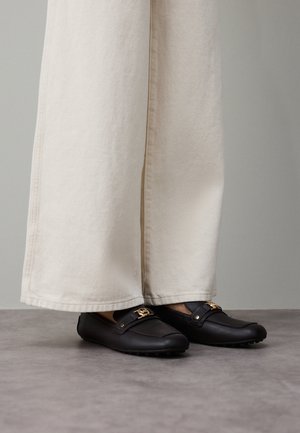Mocassini in pelle nera con dettagli in oro, indossati con pantaloni larghi color crema, che mostrano una texture liscia e una forma arrotondata.