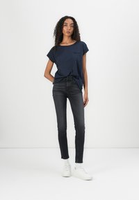 Camiseta de manga corta en azul marino con un bolsillo frontal, combinada con jeans oscuros de corte ajustado y zapatillas blancas, sobre un fondo liso.