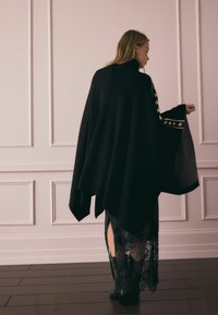 Fekete kötött poncsó magas gallérral, arany csillag díszítéssel az ujjakon; fekete, csipkés szegélyű ruha fölött viselve.