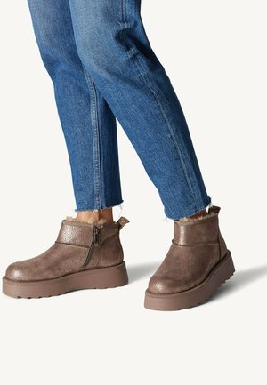 Stivali alla caviglia marrone metallico con una finitura texturizzata, zip laterale e una suola spessa a contrasto. Abbinati a jeans blu con orlo sfrangiato.