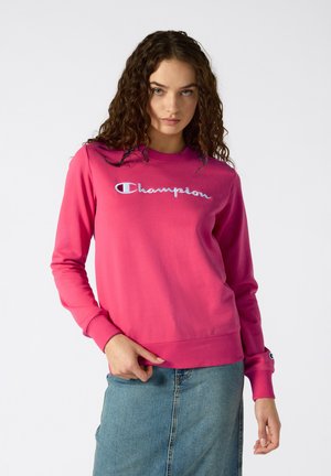 Sweatshirt rose avec manches longues, col rond et logo "Champion" brodé sur le devant. Tissu lisse, design simple.