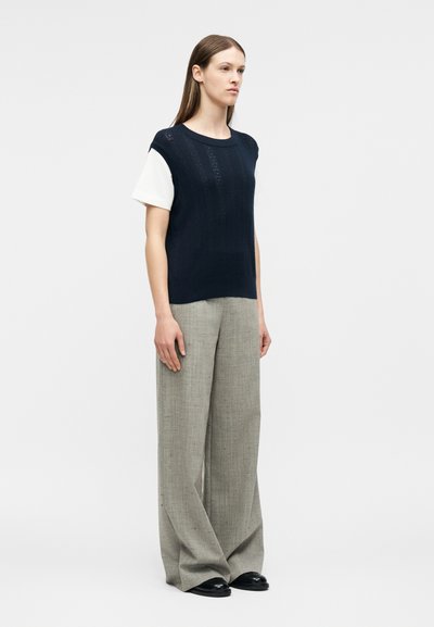 Γυναίκα που στέκεται φορώντας ένα μπλε σκουρομωβ πουλόβερ χωρίς μανίκια πάνω από λευκό κοντομάνικο, φαρδιές γκρι παντελόνες και μαύρα παπούτσια, ουδέτερο φόντο.