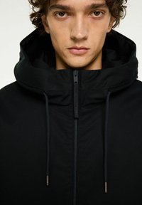 Ung person som bär en svart jacka med hög krage och huva, vänd framåt med ett neutralt uttryck mot en ljus bakgrund.