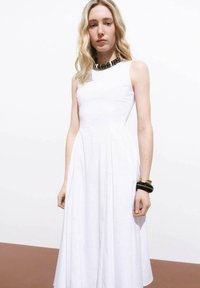Robe blanche sans manches avec une silhouette en A, texture douce et détails plissés. Les accessoires comprennent un collier foncé et des bracelets.