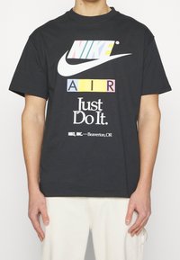 Černé bavlněné tričko s barevným logem Nike a textem "AIR" v tučných blocích, s nápisem "Just Do It" vytištěným pod ním. Krátké rukávy a volný střih.