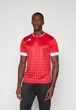 Homme noir portant un maillot de sport rouge à motif losange, bord de col blanc et pantalon noir, debout devant un fond clair uni.