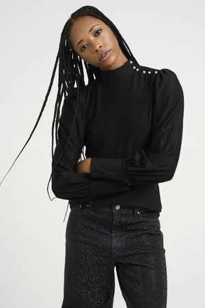 Svart långärmad topp med hög krage och puffiga axlar, prydd med dekorativa knappar på ena axeln; tillsammans med mörka, utsmyckade jeans.