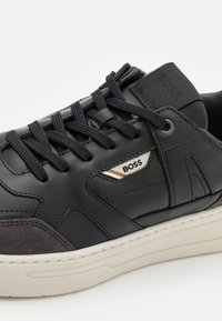 Černé kožené tenisky s šedými semišovými akcenty, texturovanou gumovou podrážkou a černými plochými tkaničkami. Obsahují decentní detail s logem "BOSS".