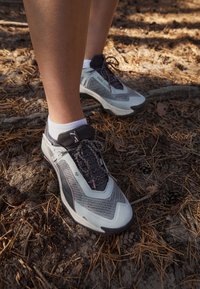 Lekki buty biegowe z siateczkową cholewką w kolorze szarym i białym, z czarnymi akcentami, teksturowaną podeszwą i elementami odblaskowymi, ustawione na naturalnej powierzchni.
