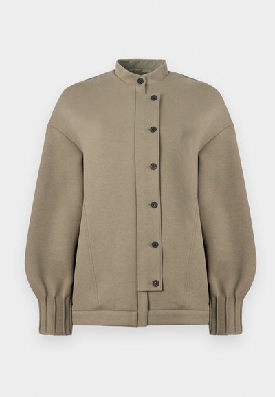 Τζάκετ σε απόχρωση ταούπ με ψηλό γιακά, ασύμμετρο κλείσιμο με κουμπιά, μακριά φουσκωτά μανίκια και πιετέ μανσέτες σε λευκό φόντο.