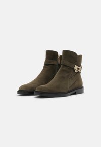 Une paire de bottines en suède vert olive avec des semelles noires, des zips sur les côtés et des détails en boucle en métal doré, sur fond neutre.