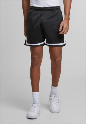 Person, die schwarze Sportshorts mit weißem Besatz, weiße Socken und weiße Reebok-Sneakers trägt, steht vor einem einfarbigen hellen Hintergrund.