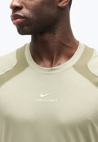 Κοντινό πλάνο ενός ατόμου που φοράει μπλουζάκι αθλητικό Nike Pro σε μπεζ χρώμα με υφασμένα πάνελ στους ώμους και μικρό λογότυπο Nike στο στήθος.