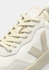 Sneaker bianca con materiale testurizzato, lacci crema e un accento logo beige. Presenta una punta arrotondata e dettagli cuciti lungo la suola.