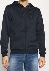 Marineblå hættetrøje med lynlås lavet af blødt stof, med snøre i hætten, to forlommer og ribstrikkede manchetter. Minimalistisk design med diskret branding.